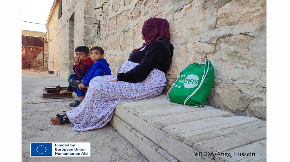 "Khaldiya sits in front of her home with her two children, her features reflecting resilience and hope for a better future after a difficult journey of displacement."